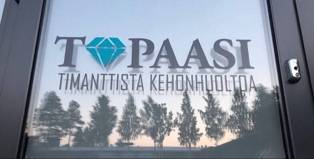 Yrityskuva Topaasin logosta lasin pinnalla. Lasiin heijastuu kauniisti vastapäätä oleva metsämaisema