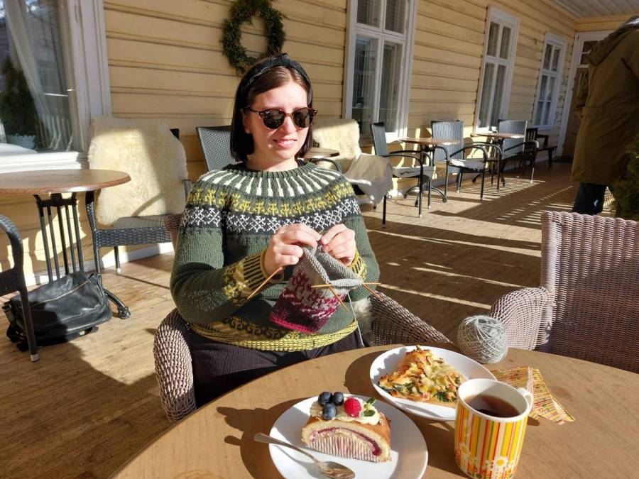 Nuori nainen neuloo aurinkoisella Mustosen talon kuistilla villapaita päällään. Edessä pöydällä on herkullinen kattaus Kauppaneuvoksen kahvilan tuotteita. 